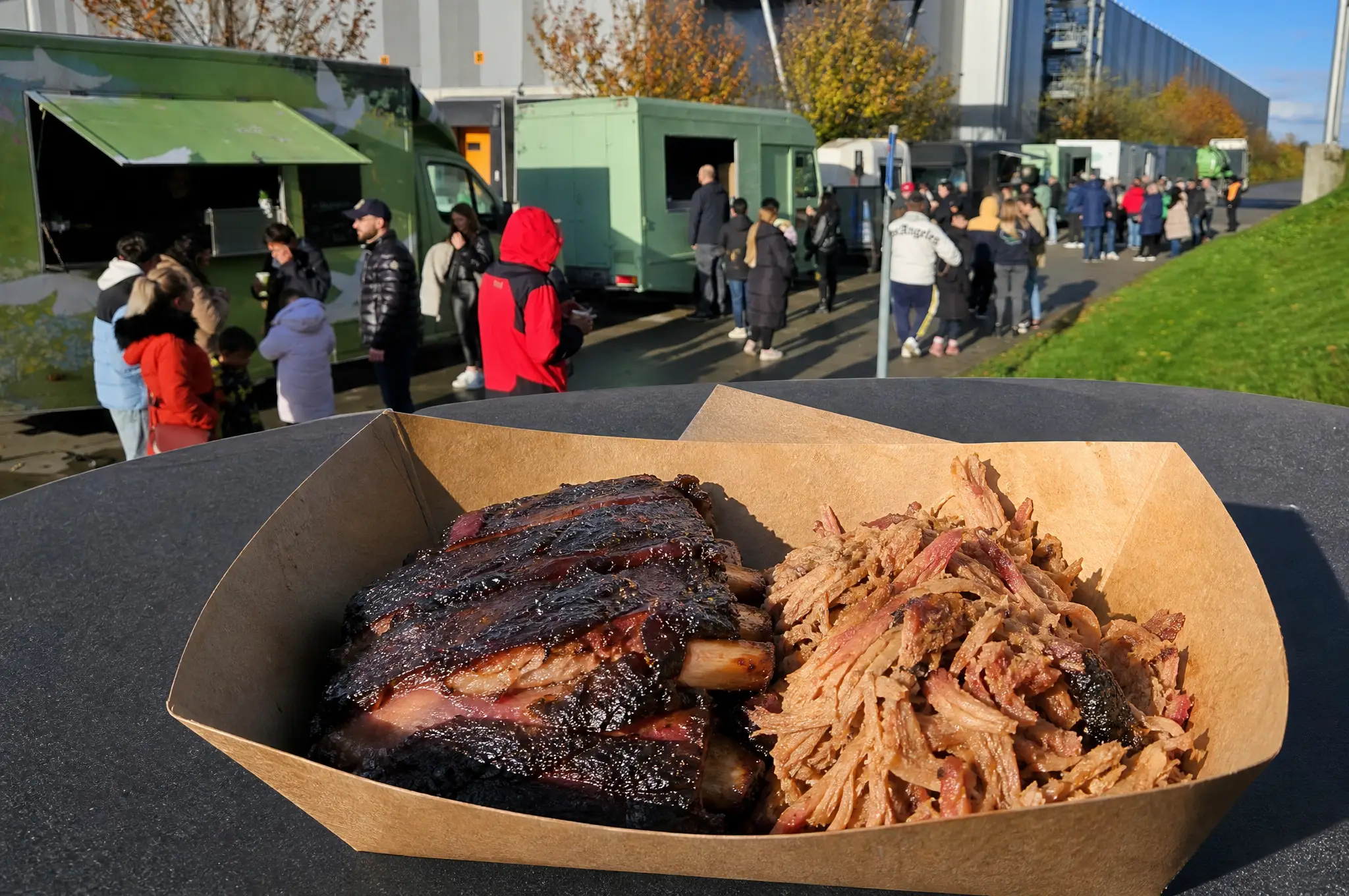 Ribs und Pulled Meat in einer Pappschale, dahinter Gäste vor grünen Foodtrucks