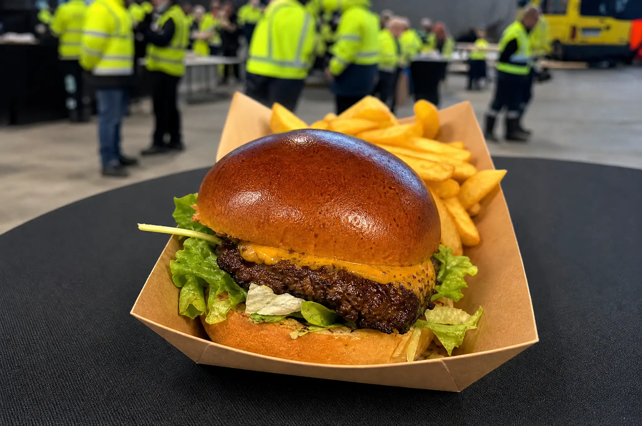 Cheeseburger mit Pommes in einer Pappschale, im Hintergrund Gäste vor den Trucks