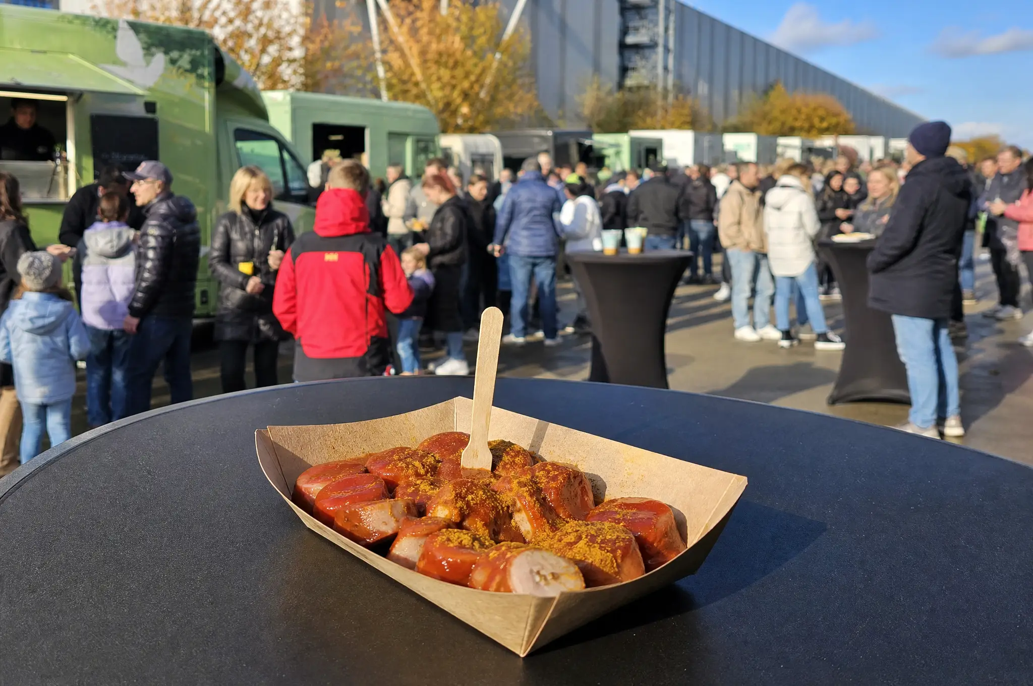 Currywurst mit Currysoße in einer Pappschale, dahinter Gäste an Stehtischen