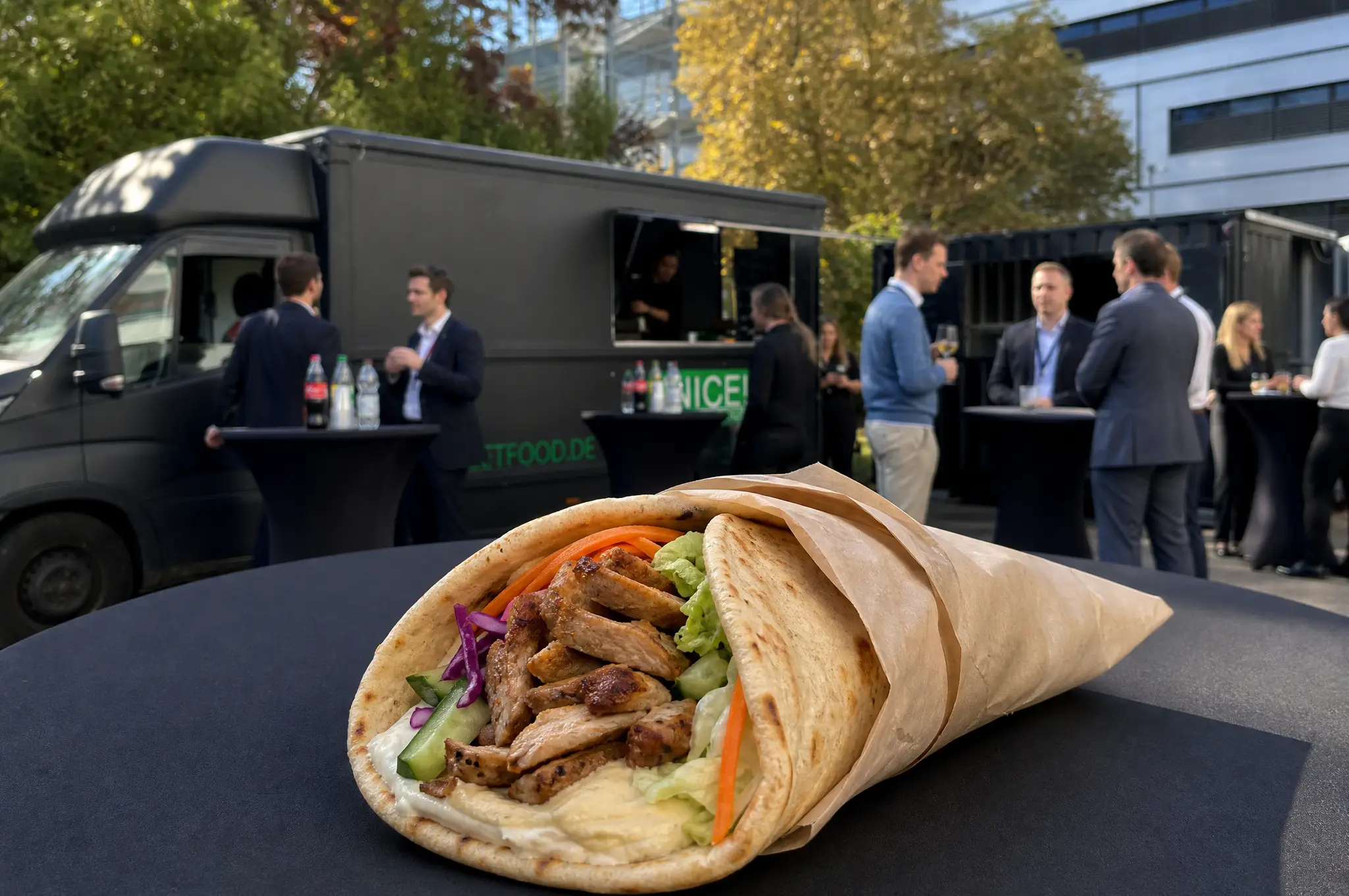 Wrap mit gegrilltem Fleisch auf einem Stehtisch, dahinter Gäste vor schwarzen Foodtrucks
