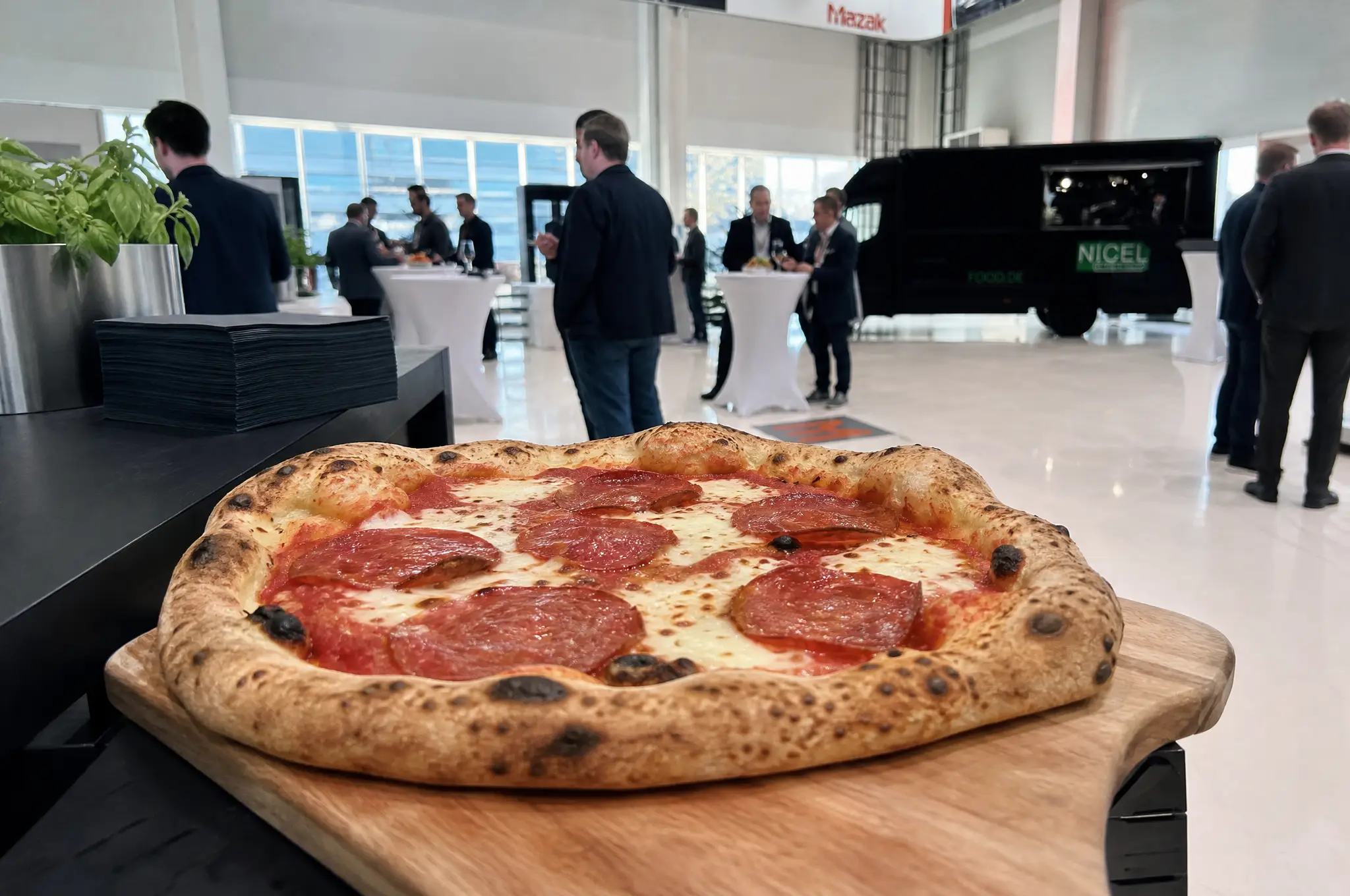 Pizza mit Salami auf einem Holzbrett, im Hintergrund eine Veranstaltung in einer Halle