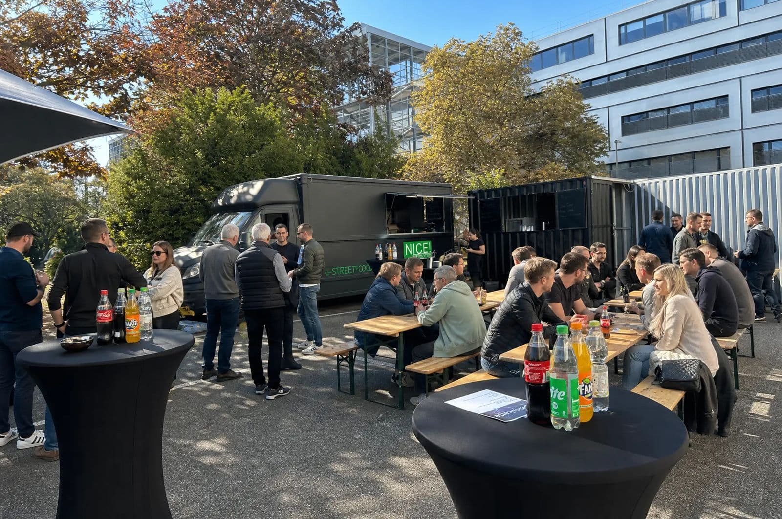 Gäste an Stehtischen und Bierbänken vor zwei NICE! Streetfood Trucks auf einem Firmengelände
